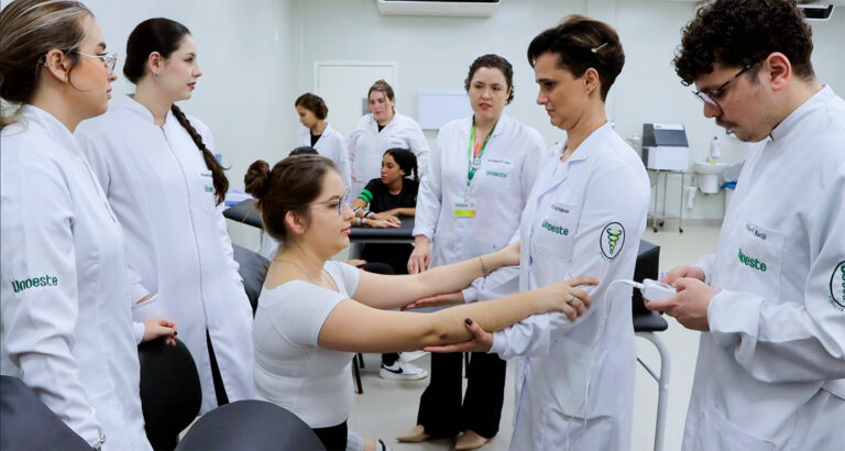 Estudantes de Fisioterapia da Unoeste em aula prática, avaliando movimentos do braço de uma colega sob supervisão de professor, em ambiente de laboratório.