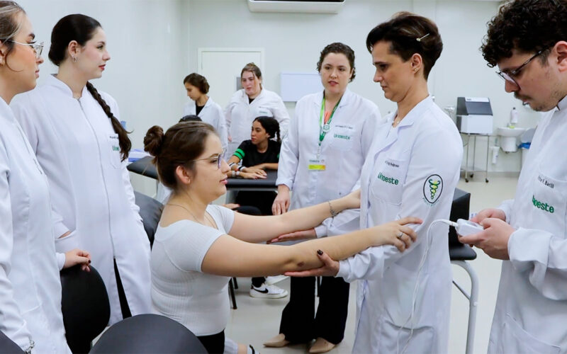 Estudantes de Fisioterapia da Unoeste em aula prática, avaliando movimentos do braço de uma colega sob supervisão de professor, em ambiente de laboratório.