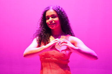Jovem sorridente com fundo iluminado em rosa, fazendo o gesto de coração com as mãos em apoio à campanha Outubro Rosa de conscientização sobre a prevenção do câncer de mama.