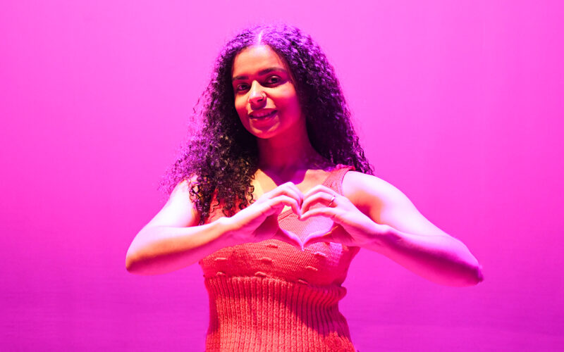 Jovem sorridente com fundo iluminado em rosa, fazendo o gesto de coração com as mãos em apoio à campanha Outubro Rosa de conscientização sobre a prevenção do câncer de mama.