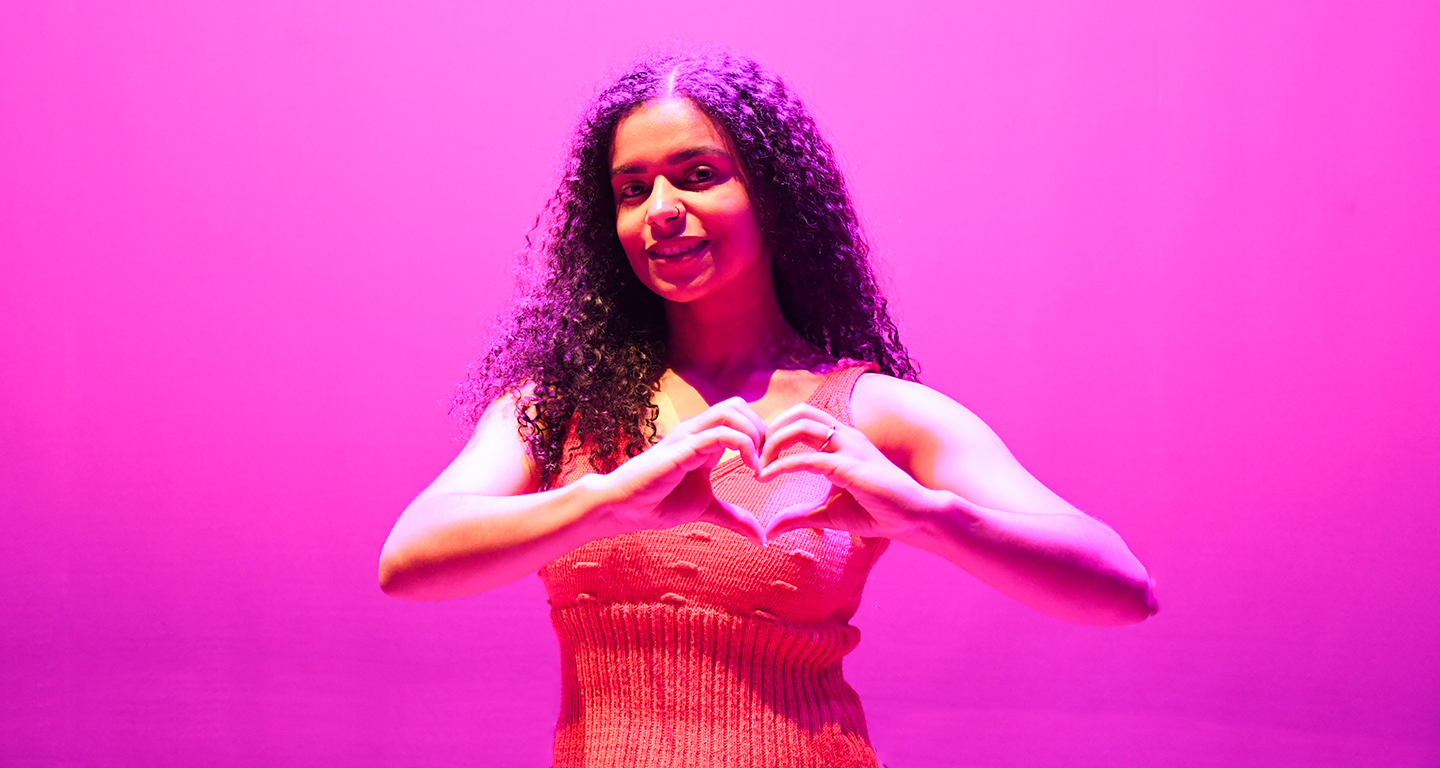 Jovem sorridente com fundo iluminado em rosa, fazendo o gesto de coração com as mãos em apoio à campanha Outubro Rosa de conscientização sobre a prevenção do câncer de mama.