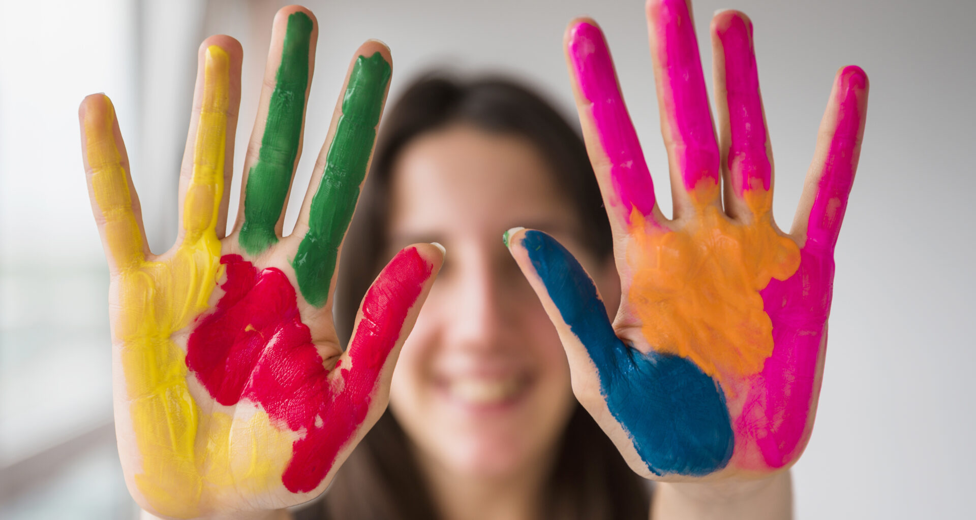Duas mãos abertas com tinta colorida nas palmas, exibindo as cores amarelo, vermelho, verde, azul, rosa e laranja, com rosto desfocado ao fundo.