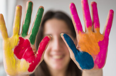 Duas mãos abertas com tinta colorida nas palmas, exibindo as cores amarelo, vermelho, verde, azul, rosa e laranja, com rosto desfocado ao fundo.