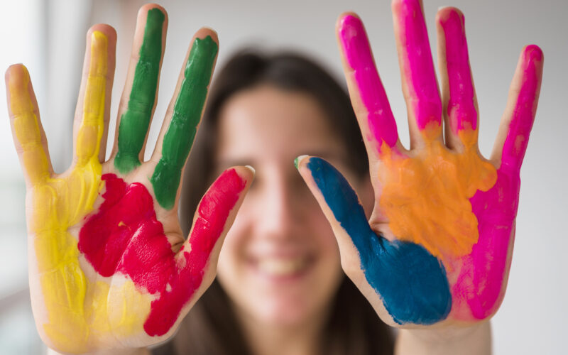 Duas mãos abertas com tinta colorida nas palmas, exibindo as cores amarelo, vermelho, verde, azul, rosa e laranja, com rosto desfocado ao fundo.