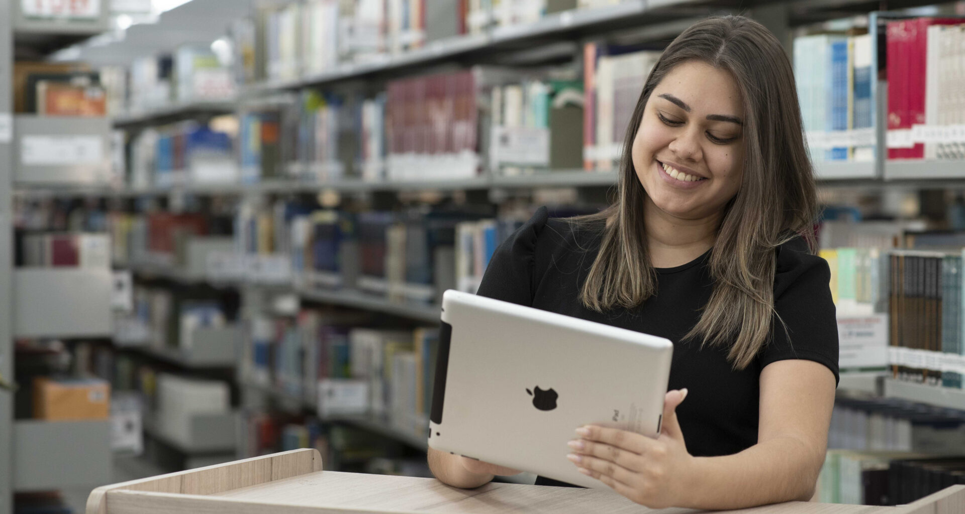 Jovem sentada em uma biblioteca universitária, usando um tablet e sorrindo, com estantes de livros ao fundo.