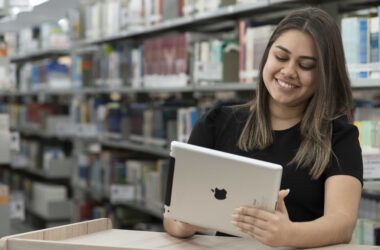 Jovem sentada em uma biblioteca universitária, usando um tablet e sorrindo, com estantes de livros ao fundo.