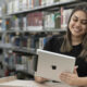 Jovem sentada em uma biblioteca universitária, usando um tablet e sorrindo, com estantes de livros ao fundo.
