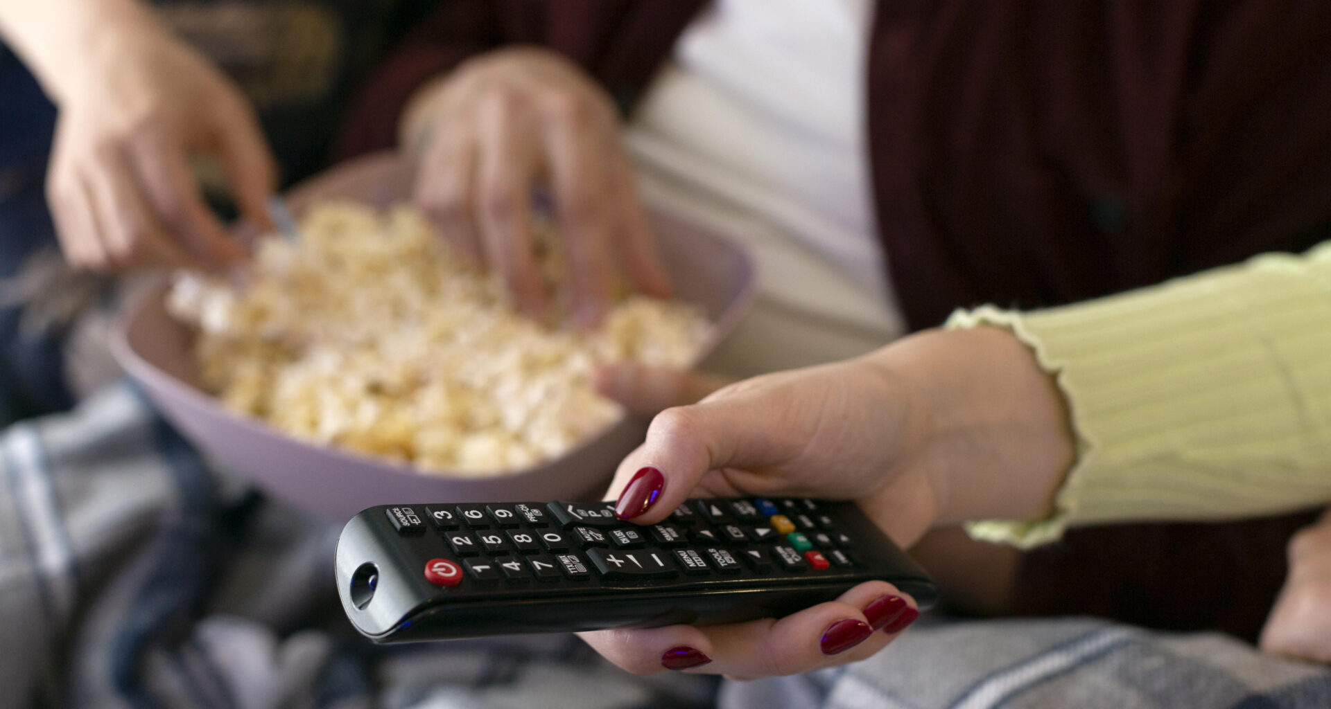 Mãos segurando controle remoto e tigela com pipoca sobre o colo, em ambiente de sala de estar, prontas para assistir à TV.