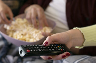 Mãos segurando controle remoto e tigela com pipoca sobre o colo, em ambiente de sala de estar, prontas para assistir à TV.