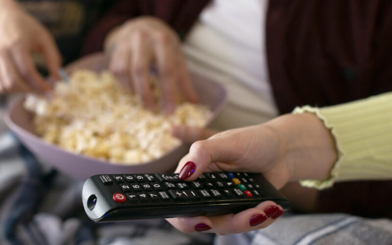 Mãos segurando controle remoto e tigela com pipoca sobre o colo, em ambiente de sala de estar, prontas para assistir à TV.