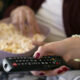 Mãos segurando controle remoto e tigela com pipoca sobre o colo, em ambiente de sala de estar, prontas para assistir à TV.