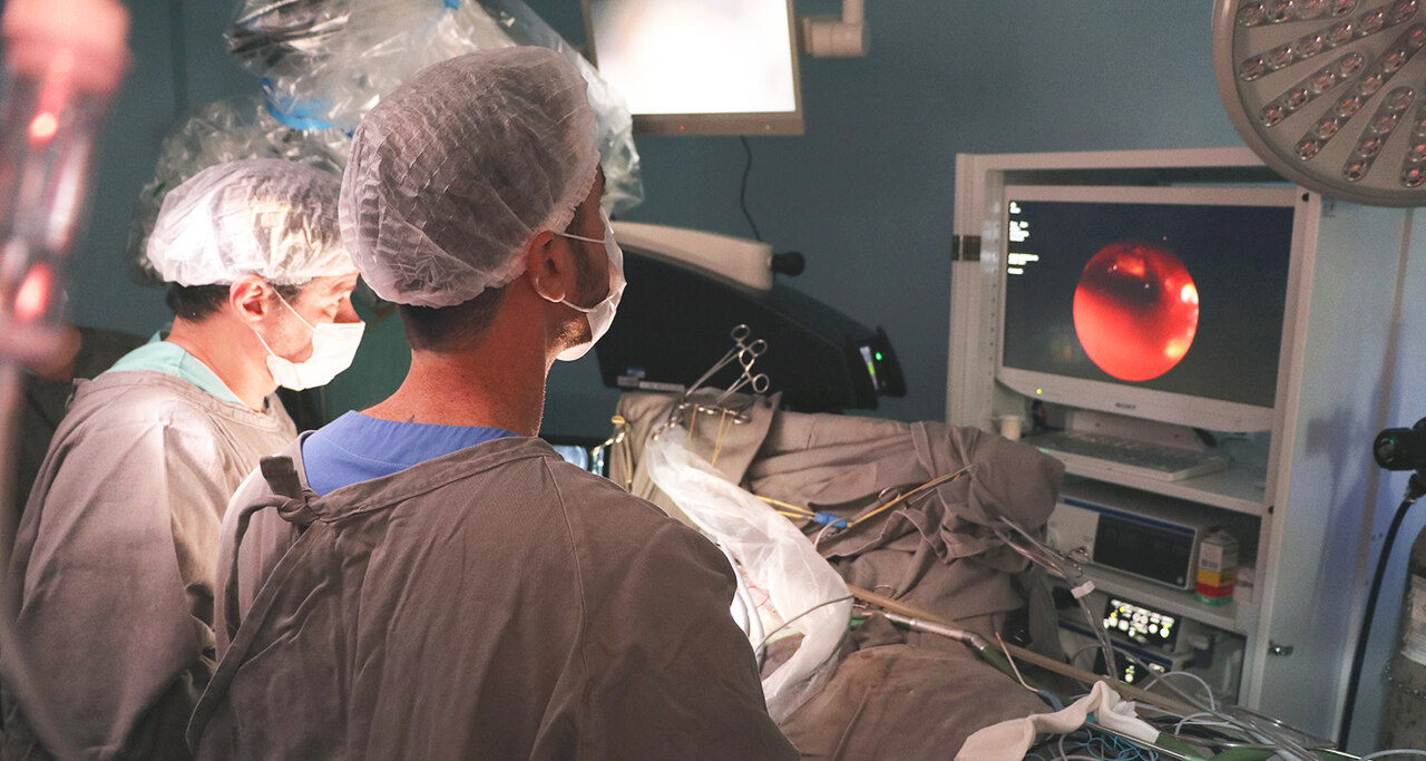 Médicos equipados com aventais estéreis e toucas cirúrgicas realizam um procedimento de alta complexidade no centro cirúrgico do Hospital de Esperança. Um dos profissionais acompanha as imagens ampliadas do interior do crânio exibidas em um monitor, enquanto instrumentos e dispositivos conectados permanecem organizados ao lado do paciente, ilustrando o uso de tecnologias avançadas na cirurgia mencionada no texto.
