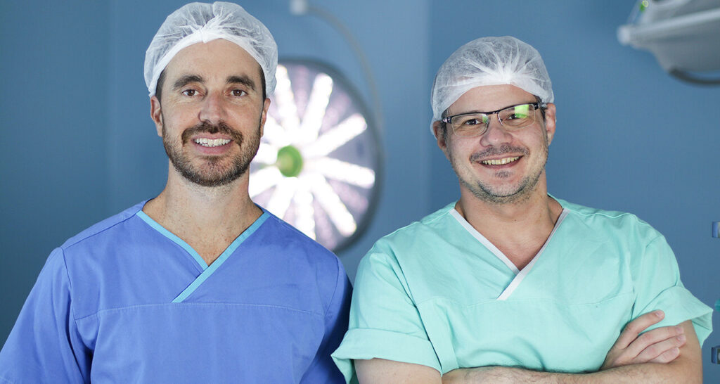  O neurocirurgião Felipe Gaia (à esquerda) e o neurocirurgião Marcio Rassi (à direita) posam sorrindo em uma sala de cirurgia, ambos usando aventais e toucas descartáveis. Ao fundo, um foco cirúrgico desfocado reforça o ambiente hospitalar relacionado ao procedimento mencionado no texto.