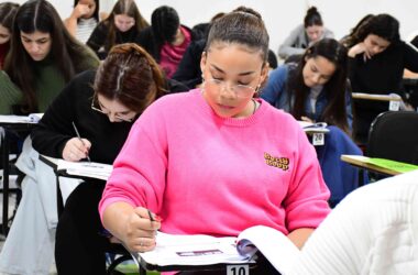 Estudantes realizando prova em sala de aula, concentrados em suas folhas de resposta; em destaque, uma aluna com moletom rosa escreve atentamente.