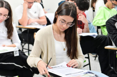 Estudantes sentados em carteiras individuais realizam uma prova escrita em sala de aula. Em primeiro plano, uma aluna concentra-se na leitura e resolução das questões, enquanto outros candidatos respondem à avaliação ao fundo, ilustrando o ingresso na faculdade por meio da nota do Enem e de outros processos seletivos.
