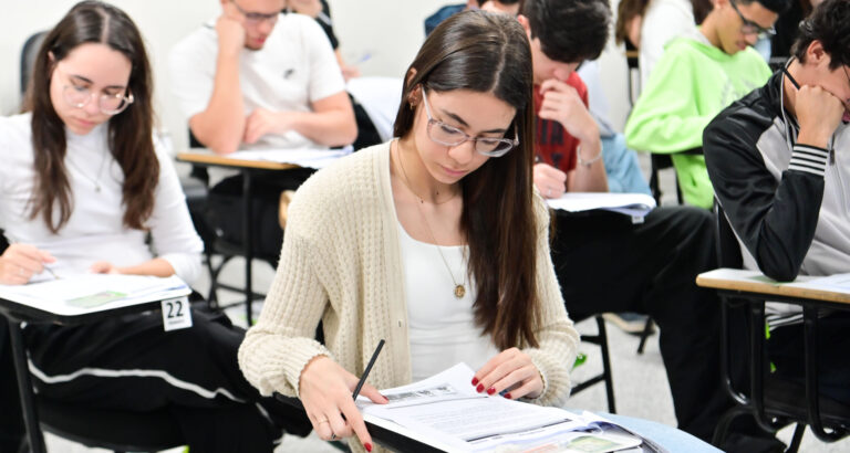 Estudantes sentados em carteiras individuais realizam uma prova escrita em sala de aula. Em primeiro plano, uma aluna concentra-se na leitura e resolução das questões, enquanto outros candidatos respondem à avaliação ao fundo, ilustrando o ingresso na faculdade por meio da nota do Enem e de outros processos seletivos.