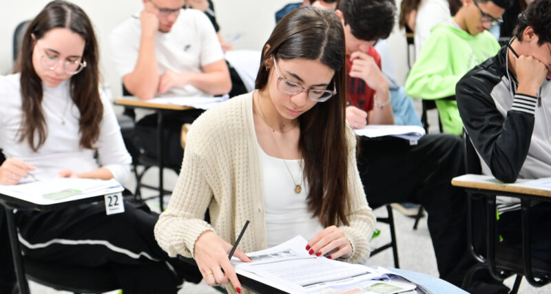 Estudantes sentados em carteiras individuais realizam uma prova escrita em sala de aula. Em primeiro plano, uma aluna concentra-se na leitura e resolução das questões, enquanto outros candidatos respondem à avaliação ao fundo, ilustrando o ingresso na faculdade por meio da nota do Enem e de outros processos seletivos.