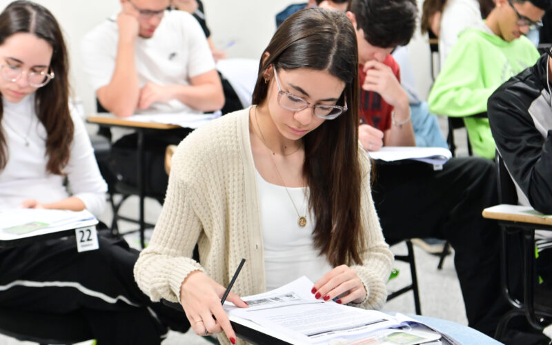 Estudantes sentados em carteiras individuais realizam uma prova escrita em sala de aula. Em primeiro plano, uma aluna concentra-se na leitura e resolução das questões, enquanto outros candidatos respondem à avaliação ao fundo, ilustrando o ingresso na faculdade por meio da nota do Enem e de outros processos seletivos.