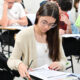 Estudantes sentados em carteiras individuais realizam uma prova escrita em sala de aula. Em primeiro plano, uma aluna concentra-se na leitura e resolução das questões, enquanto outros candidatos respondem à avaliação ao fundo, ilustrando o ingresso na faculdade por meio da nota do Enem e de outros processos seletivos.