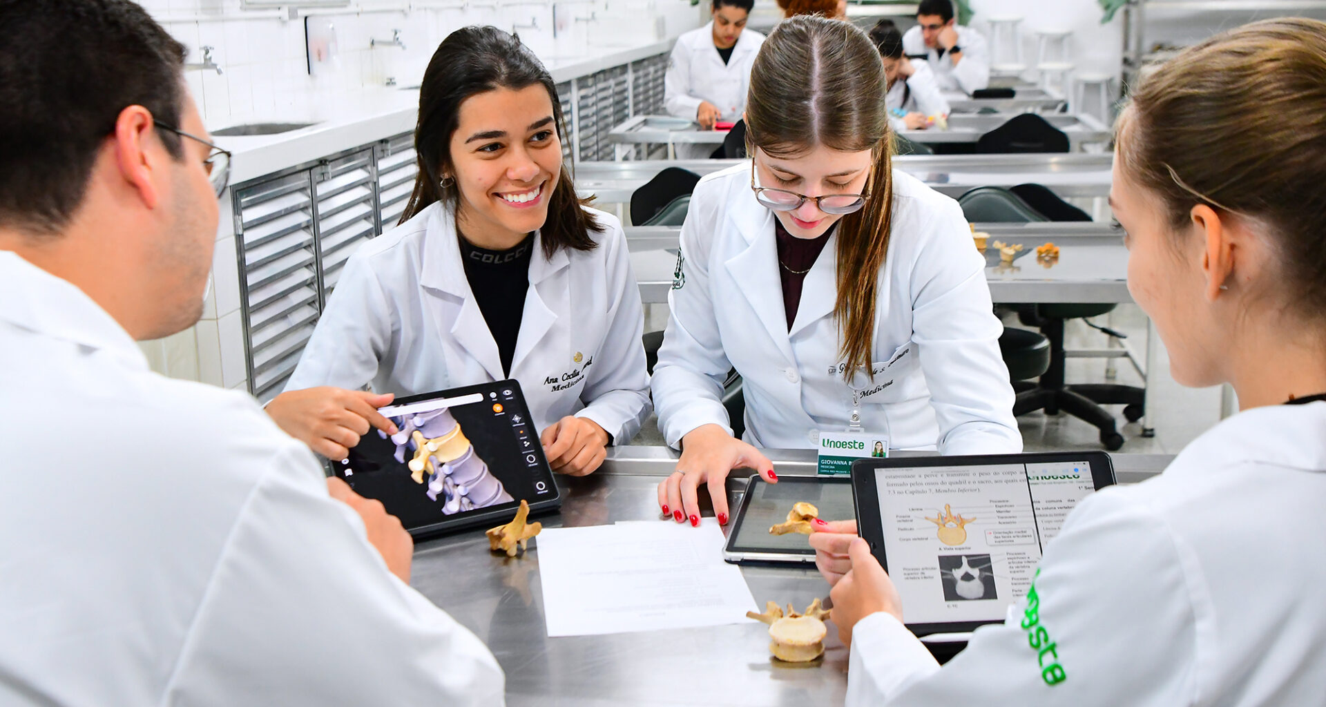 Alunos da graduação em Medicina da Unoeste, estudam anatomia em laboratório com jalecos brancos, utilizando tablets e modelos de vértebras humanas para aulas práticas do curso de Medicina.