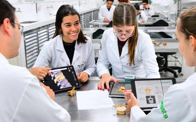 Alunos da graduação em Medicina da Unoeste, estudam anatomia em laboratório com jalecos brancos, utilizando tablets e modelos de vértebras humanas para aulas práticas do curso de Medicina.