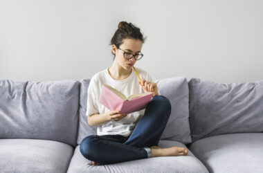 Jovem sentada em um sofá cinza, lendo um caderno rosa e segurando um lápis enquanto estuda.