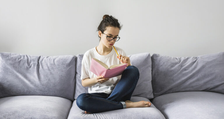 Jovem sentada em um sofá cinza, lendo um caderno rosa e segurando um lápis enquanto estuda.