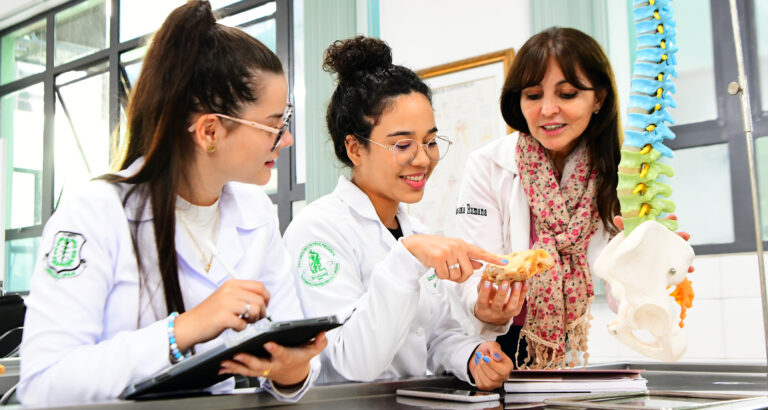 Estudantes de Medicina da Unoeste em aula prática de anatomia, observando uma estrutura óssea com orientação de professora. Todos usam jaleco branco e estão em ambiente de laboratório, com modelos anatômicos e materiais de estudo sobre a bancada.