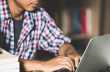 Estudante digitando em um notebook em uma biblioteca, cercado por pilhas de livros, simbolizando preparação para vestibular online.