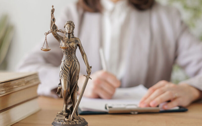 Estátua da deusa da justiça com balança e espada sobre mesa de madeira, ao lado de livros, com mulher escrevendo em prancheta ao fundo.