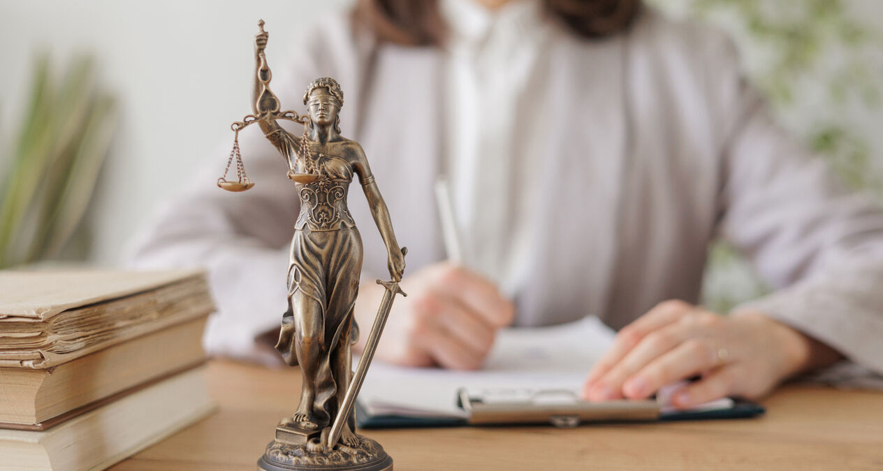 Estátua da deusa da justiça com balança e espada sobre mesa de madeira, ao lado de livros, com mulher escrevendo em prancheta ao fundo.