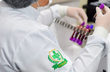 Profissional da área da saúde manuseando amostras de sangue em laboratório, com destaque para o brasão bordado no jaleco com o símbolo da Biomedicina: cruz verde, microscópio e ramos laureados.