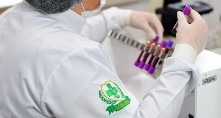 Profissional da área da saúde manuseando amostras de sangue em laboratório, com destaque para o brasão bordado no jaleco com o símbolo da Biomedicina: cruz verde, microscópio e ramos laureados.