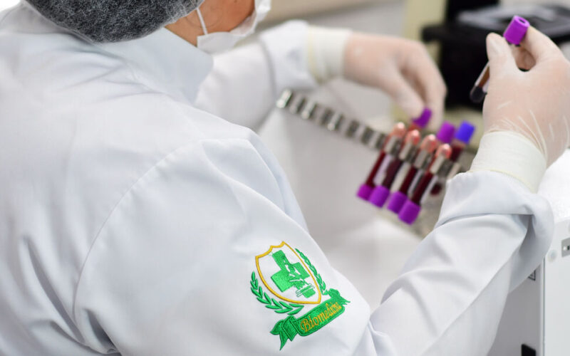 Profissional da área da saúde manuseando amostras de sangue em laboratório, com destaque para o brasão bordado no jaleco com o símbolo da Biomedicina: cruz verde, microscópio e ramos laureados.