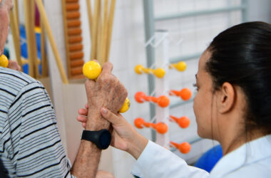 Fisioterapeuta auxilia idoso durante exercício de reabilitação com halteres amarelos em ambiente clínico, demonstrando cuidado e suporte na fisioterapia.
