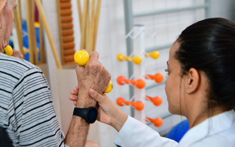 Fisioterapeuta auxilia idoso durante exercício de reabilitação com halteres amarelos em ambiente clínico, demonstrando cuidado e suporte na fisioterapia.