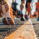 Detalhe dos pés de vários corredores usando tênis esportivos durante uma corrida de rua, com foco no movimento sobre o asfalto e outras pessoas correndo ao fundo em ambiente urbano ao ar livre.