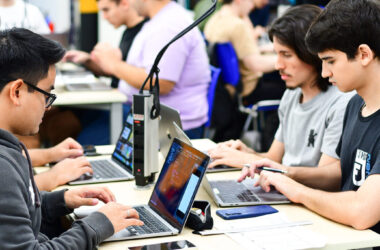 Alunos da Faculdade de Informática em aula prática, sentados lado a lado em mesas compartilhadas, utilizando laptops enquanto realizam atividades no computador em uma sala de aula moderna.