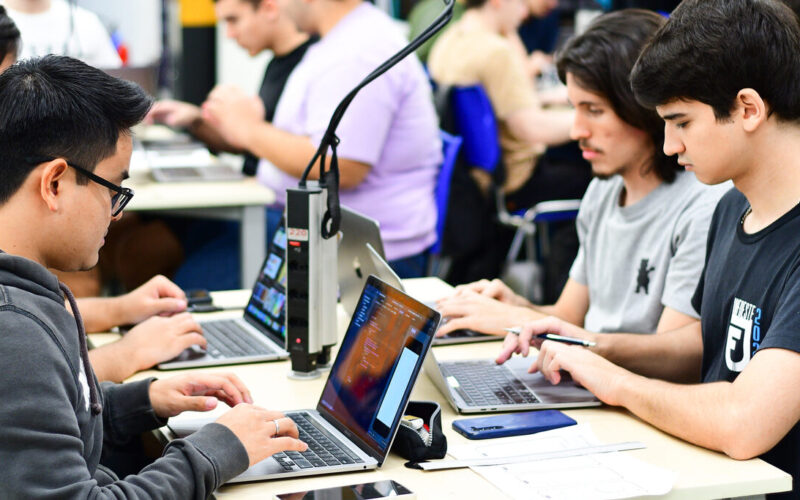 Alunos da Faculdade de Informática em aula prática, sentados lado a lado em mesas compartilhadas, utilizando laptops enquanto realizam atividades no computador em uma sala de aula moderna.
