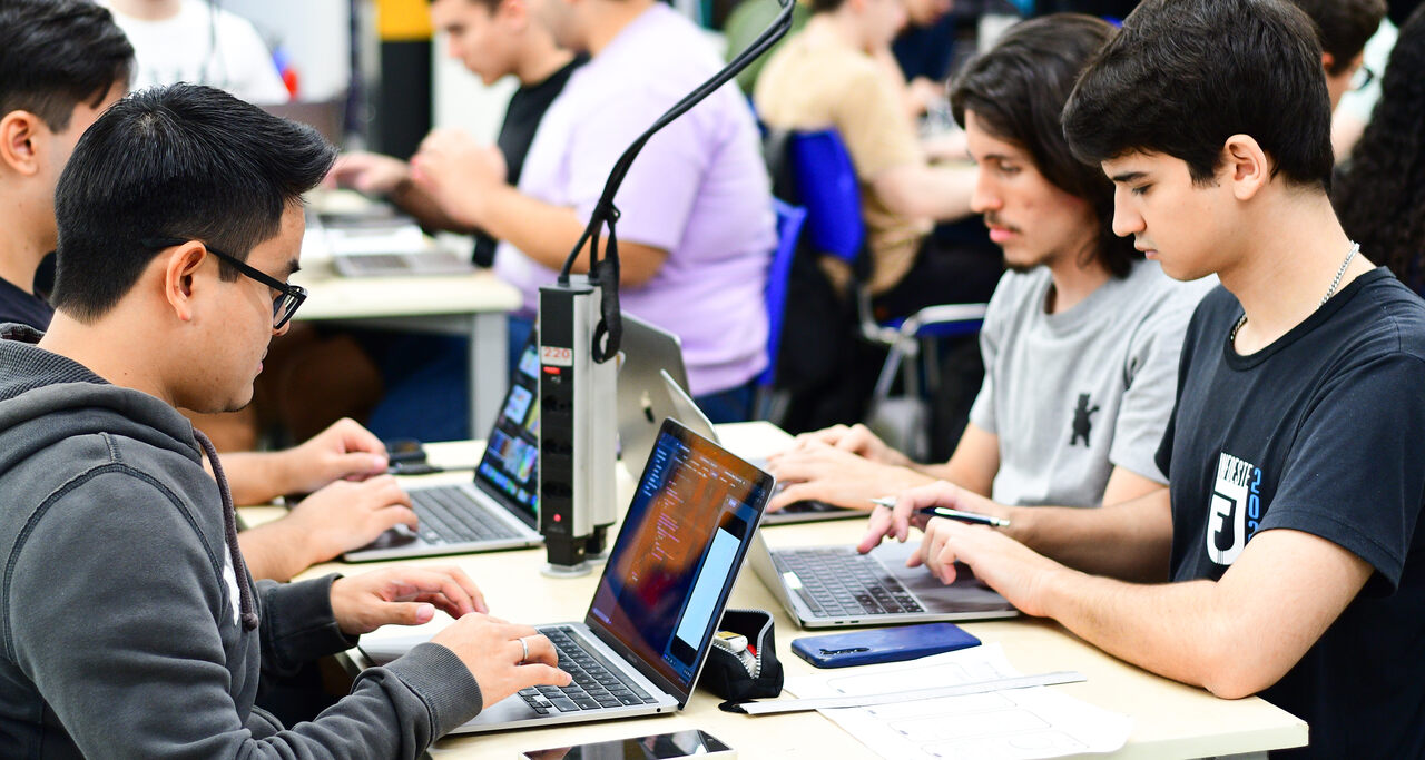 Alunos da Faculdade de Informática em aula prática, sentados lado a lado em mesas compartilhadas, utilizando laptops enquanto realizam atividades no computador em uma sala de aula moderna.