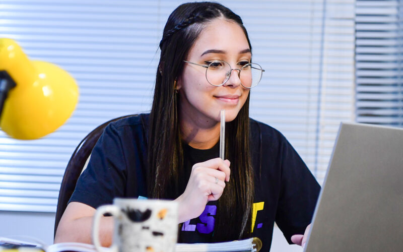Estudante sentada à mesa, usando óculos, concentrada enquanto estuda em frente ao notebook, com caderno aberto, caneca e luminária ao lado, representando a rotina de quem estuda para entrar na faculdade ou organiza os estudos on-line