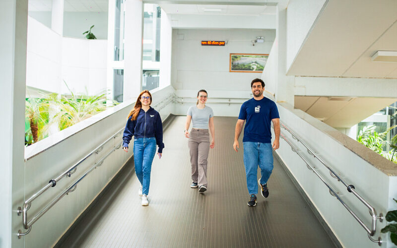 Três estudantes caminhando por uma passarela interna no campus da Unoeste, em um ambiente amplo, iluminado e rodeado por áreas verdes, representando a rotina universitária e a integração entre os espaços da instituição.