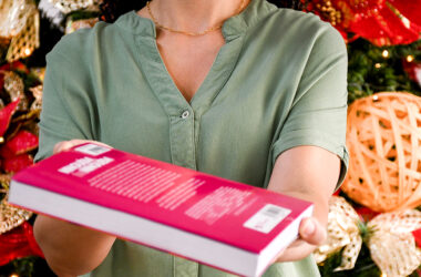 Mulher vestindo camisa verde segurando e oferecendo um livro rosa de capa rígida em frente a uma árvore de Natal decorada com laços e enfeites dourados, simbolizando o ato de presentear com livros nas festas de fim de ano.