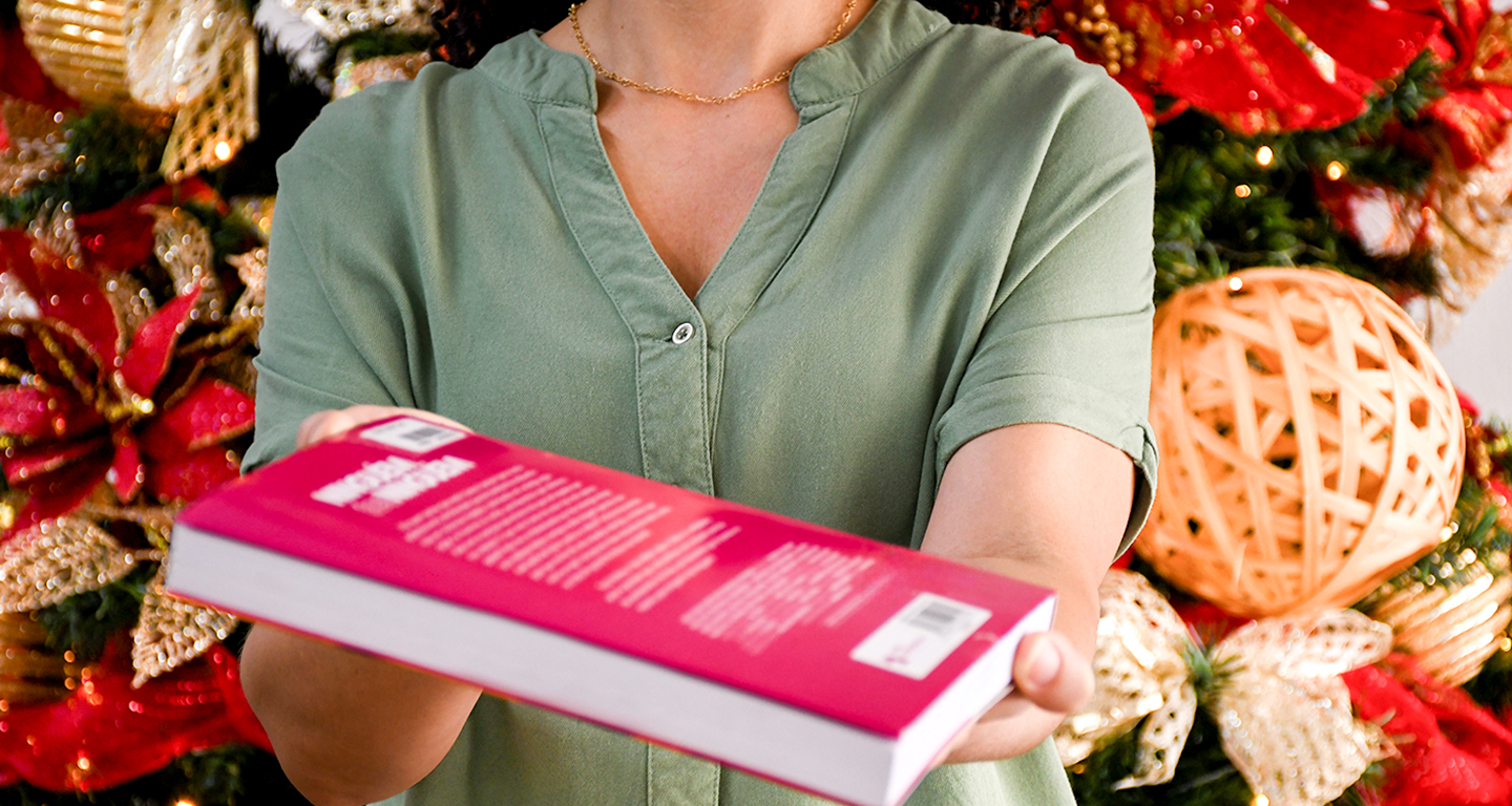Mulher vestindo camisa verde segurando e oferecendo um livro rosa de capa rígida em frente a uma árvore de Natal decorada com laços e enfeites dourados, simbolizando o ato de presentear com livros nas festas de fim de ano.