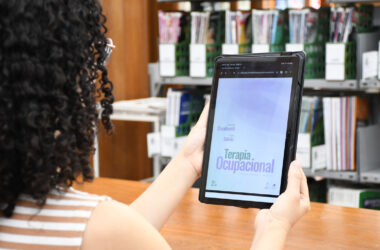 Estudante com cabelos cacheados segurando um tablet em uma biblioteca, visualizando a capa digital de um livro de Terapia Ocupacional, com prateleiras de livros e periódicos ao fundo.