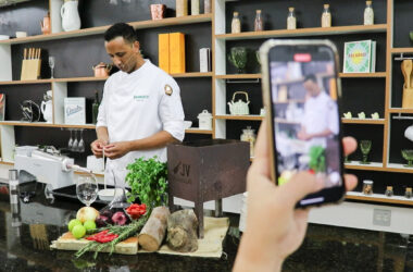 Chef usando doma branca, prepara ingredientes em uma cozinha pedagógica equipada. Ele está em frente a uma bancada com frutas, legumes, ervas e utensílios culinários, enquanto uma pessoa segura um celular filmando a cena. Ao fundo, há prateleiras com livros, potes de vidro, louças e objetos de cozinha, reforçando o ambiente de aula prática do curso de Gastronomia da Unoeste.