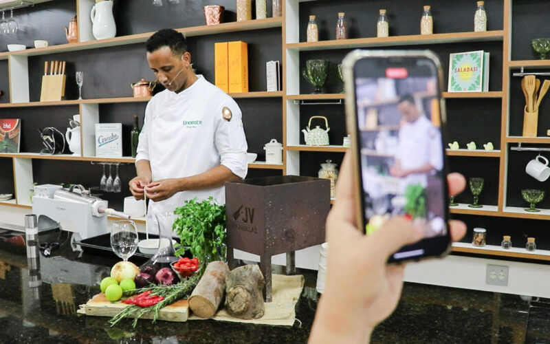 Chef usando doma branca, prepara ingredientes em uma cozinha pedagógica equipada. Ele está em frente a uma bancada com frutas, legumes, ervas e utensílios culinários, enquanto uma pessoa segura um celular filmando a cena. Ao fundo, há prateleiras com livros, potes de vidro, louças e objetos de cozinha, reforçando o ambiente de aula prática do curso de Gastronomia da Unoeste.