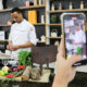 Chef usando doma branca, prepara ingredientes em uma cozinha pedagógica equipada. Ele está em frente a uma bancada com frutas, legumes, ervas e utensílios culinários, enquanto uma pessoa segura um celular filmando a cena. Ao fundo, há prateleiras com livros, potes de vidro, louças e objetos de cozinha, reforçando o ambiente de aula prática do curso de Gastronomia da Unoeste.