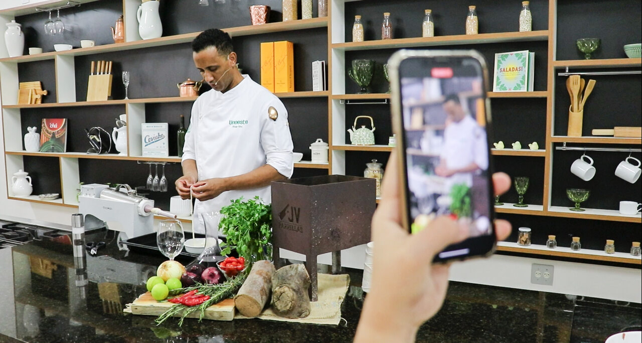 Chef usando doma branca, prepara ingredientes em uma cozinha pedagógica equipada. Ele está em frente a uma bancada com frutas, legumes, ervas e utensílios culinários, enquanto uma pessoa segura um celular filmando a cena. Ao fundo, há prateleiras com livros, potes de vidro, louças e objetos de cozinha, reforçando o ambiente de aula prática do curso de Gastronomia da Unoeste.