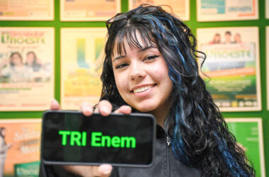 Estudante segura um celular com a frase “TRI Enem” exibida na tela, em frente a um painel de cartazes acadêmicos.